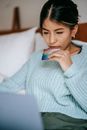 Crop concentrated Asian female wearing blue sweater making online payment while browsing netbook and lying on bed with credit card