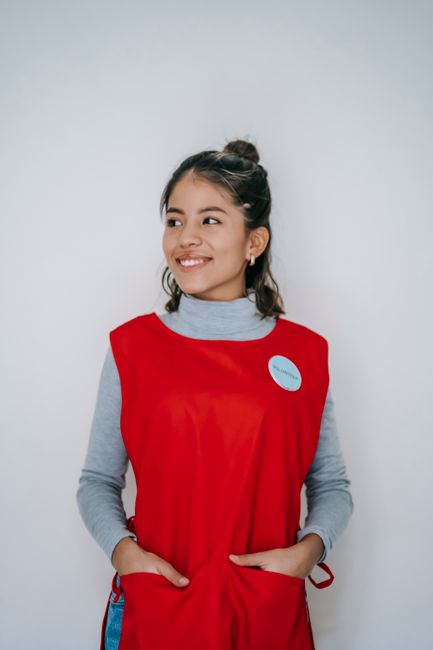 Positive young female in vivid red apron smiling and looking away on white background
