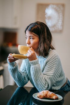 A young woman enjoys a warm coffee with a croissant in a cozy indoor setting.