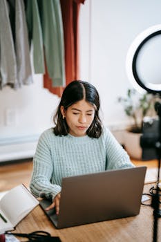 Focused young woman using laptop at home office with casual attire and bright lighting.