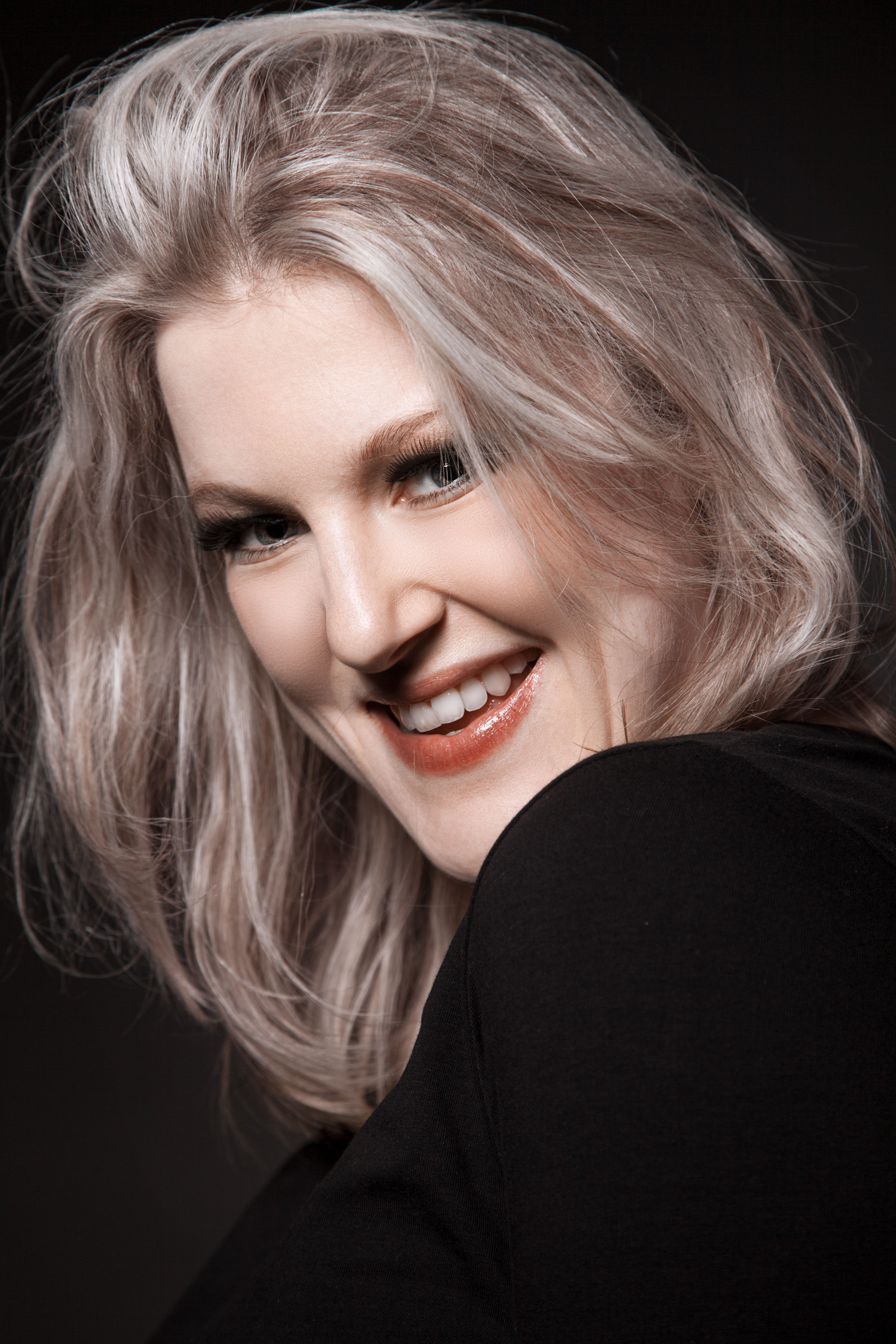 Close-up portrait of a smiling woman with blonde hair and red lips against a dark background.