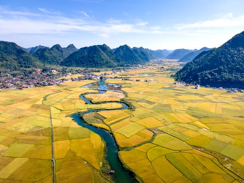 Free Drone view of curved river between green plantations with houses against high green mounts under cloudy sky in sunlight Stock Photo