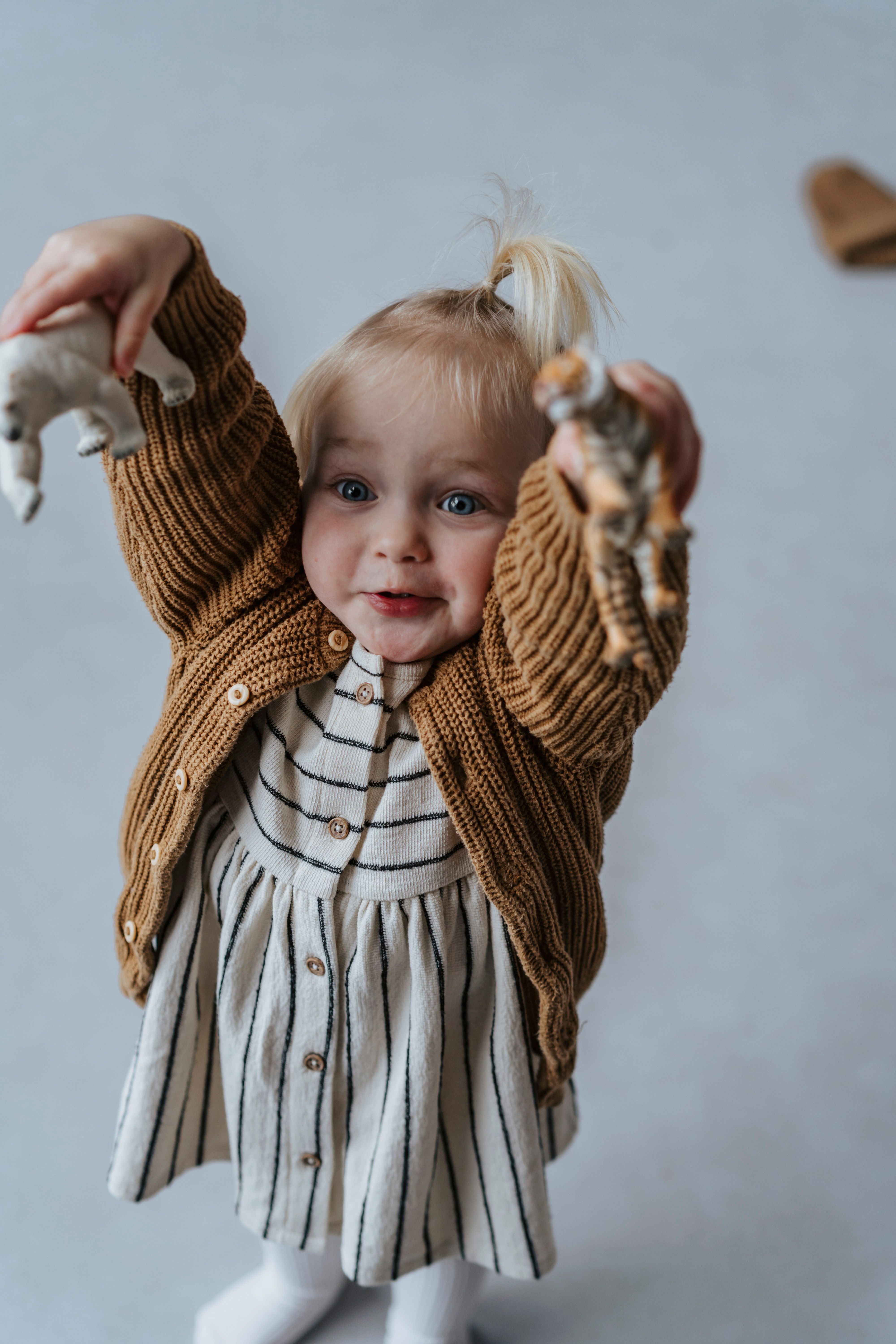 Adorable blonde girl excitedly raises animal toys indoors, showcasing joy and playfulness.