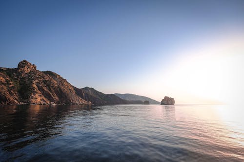 Bright sun shining in blue sky over sea surrounded by rocky hills and reflecting in rippling water