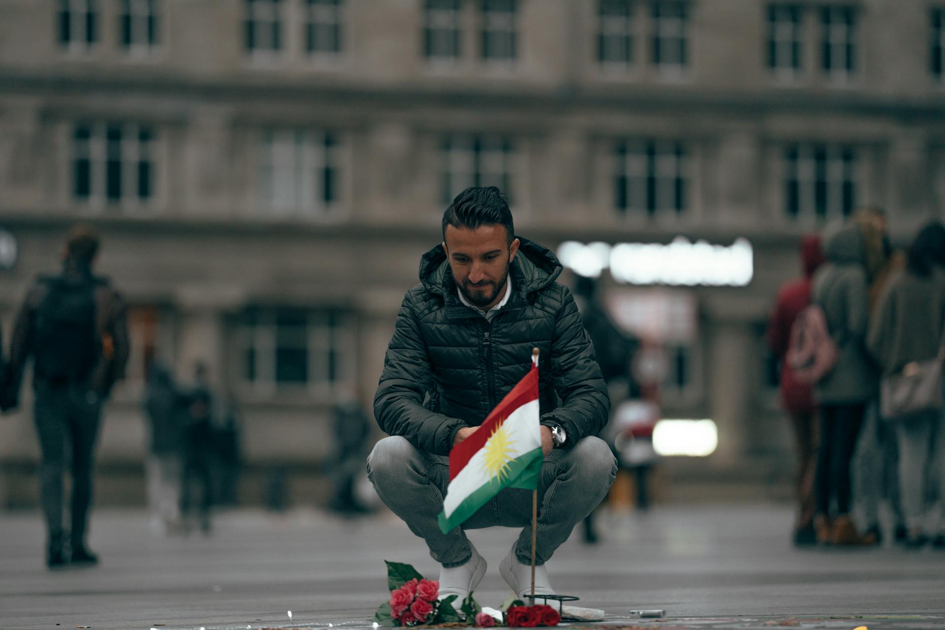 A man crouches in a public square holding a Kurdish flag, symbolizing remembrance.