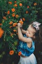 Side view of adorable capricious little girl in casual outfit with bow picking red blossoming flowers