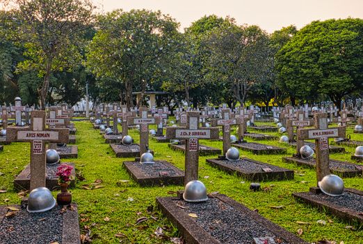 Various tombstones with hardhats and flowers in vase located in national main heroes cemetery with tall green trees in Kalibata