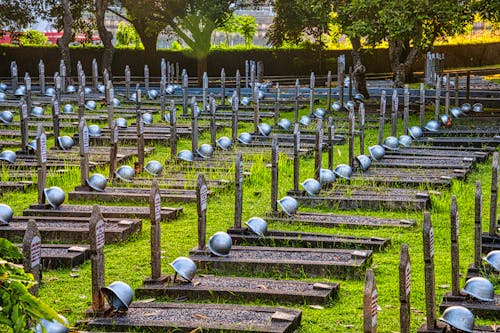 Tombstones with military hardhats located on grassy ground near tall green tall trees in national main heroes cemetery in Indonesia