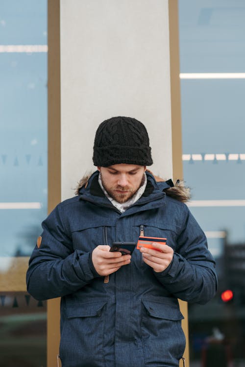 Serious young guy in warm clothes and hat standing on city street and browsing smartphone while making online payment with credit card near modern building