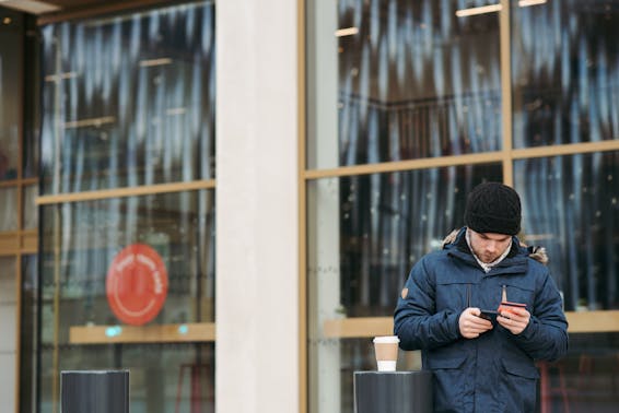 Focused young male in warm outerwear and hat standing near modern building with takeaway coffee and using smartphone while making online shopping with credit card