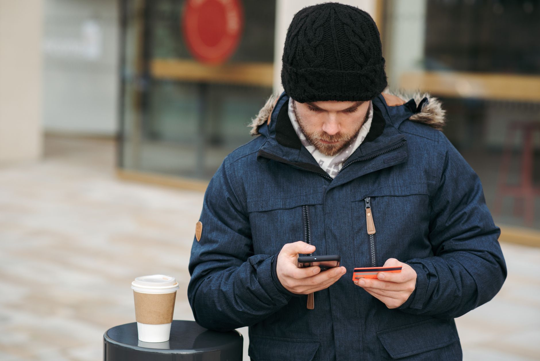Concentrated young man in warm clothes and hat entering details of credit card while making online purchase via smartphone standing on city street with takeaway coffee