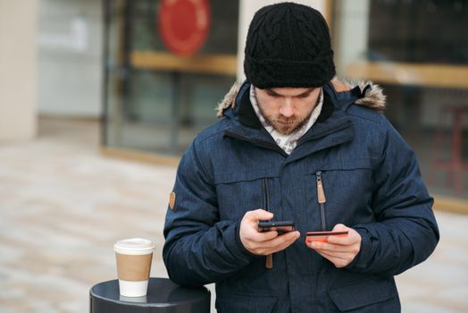 Concentrated young man in warm clothes and hat entering details of credit card while making online purchase via smartphone standing on city street with takeaway coffee