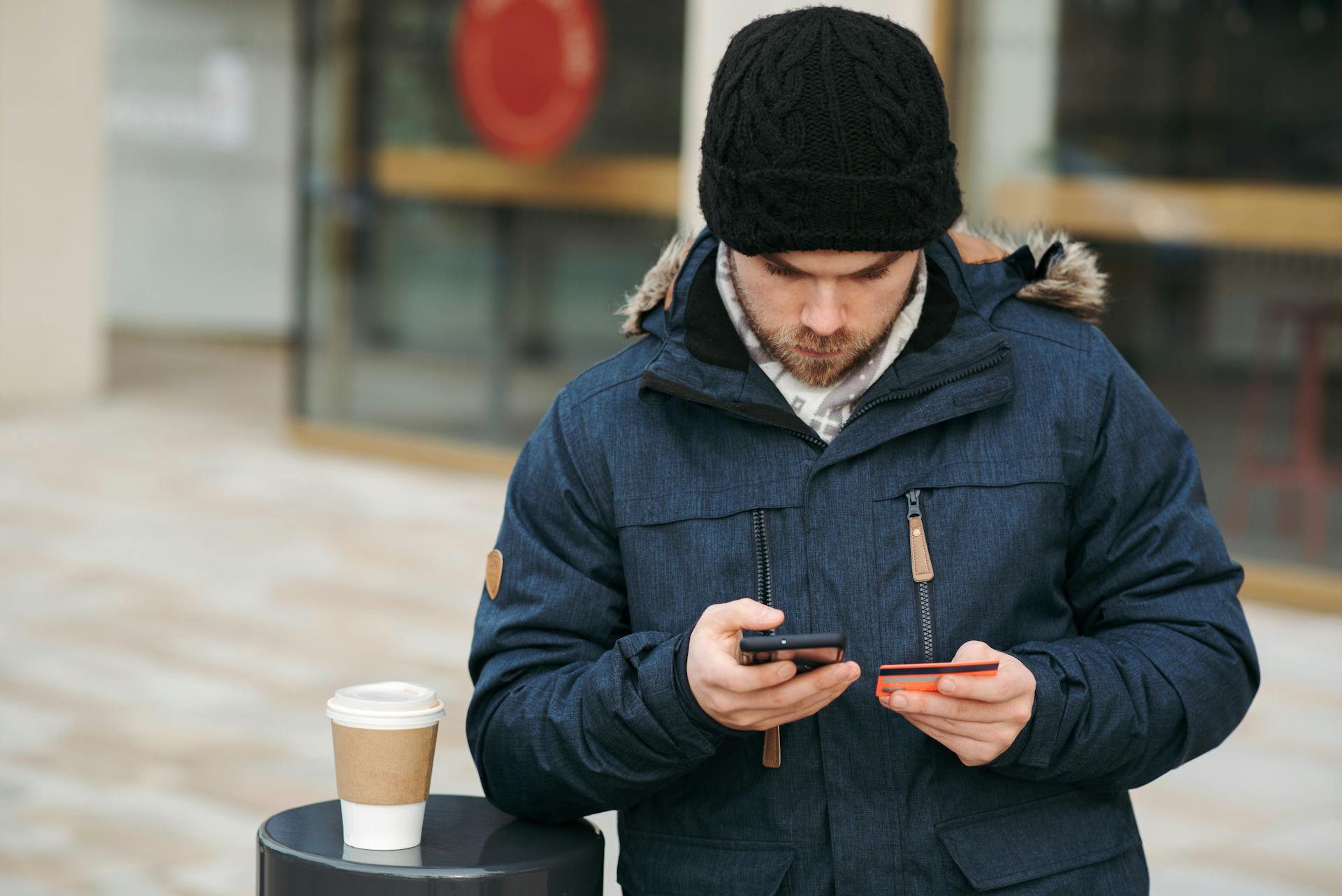 Man using smartphone and credit card for online purchase outdoors, with coffee cup nearby.