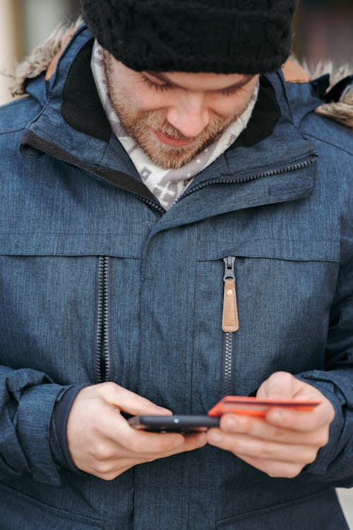Crop positive young male in stylish warm clothes and hat checking transaction details while standing on street with smartphone and credit card in hands