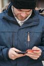 Crop positive young male in stylish warm clothes and hat checking transaction details while standing on street with smartphone and credit card in hands