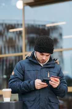 Calm young male millennial in warm outerwear and hat standing on city street with smartphone and credit card and drinking takeaway coffee while paying online for order