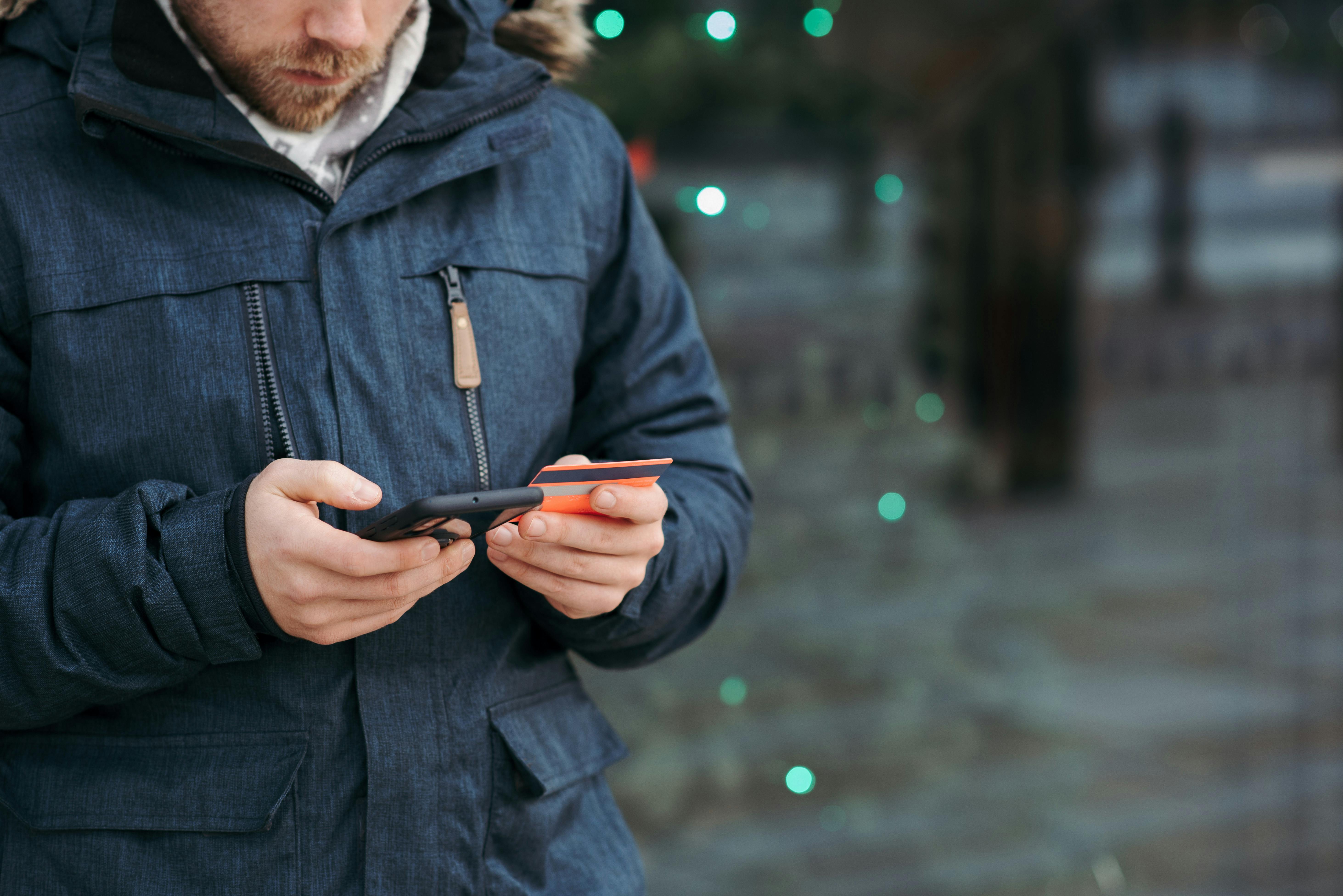 man browsing smartphone and holding credit card on street