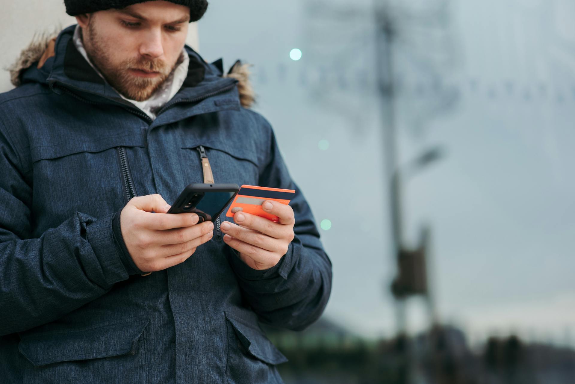 Crop concentrated man in warm clothes entering credentials of credit card on mobile phone while standing in street in daytime