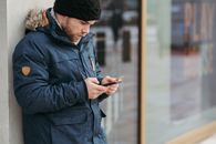 Serious man leaning on wall of building and using smartphone on street