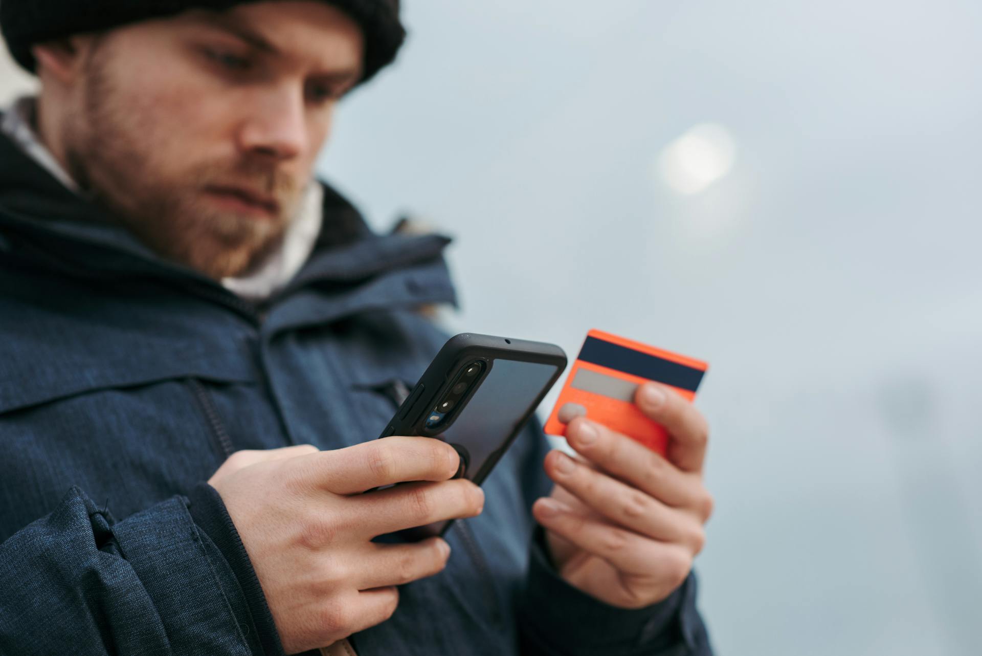 A man wearing a winter jacket uses a smartphone and credit card for an online purchase.