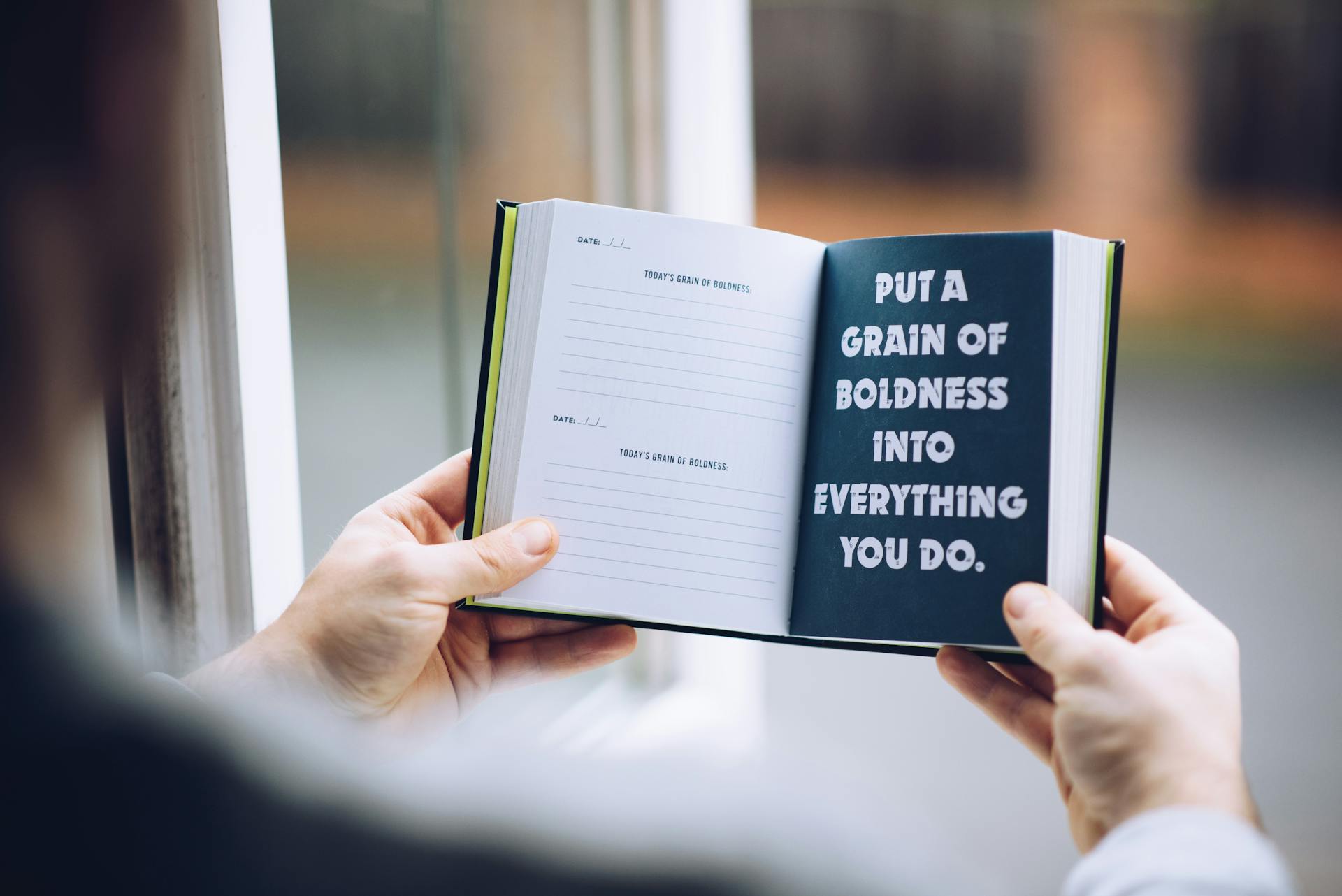 A person holds an open journal with a motivational quote by a window.