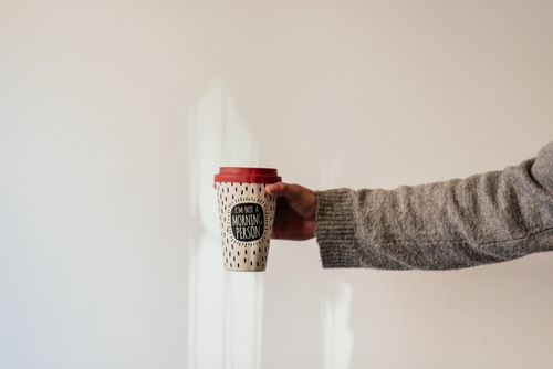 Free Crop anonymous person in soft wear demonstrating paper cup of takeaway hot drink with ornament and inscription Stock Photo