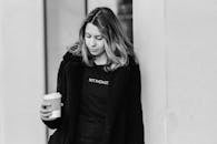 Black and white of adult tender female in outerwear with disposable glass of hot drink looking down in city