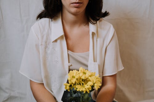 Crop unemotional brunette in white clothes with yellow gentle chrysanthemum flowers sitting against white background in light room