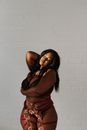 Peaceful African American female in stylish underwear revealing plump figure standing in sunlight against white brick wall