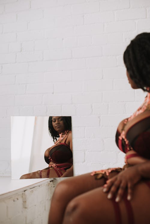 Free Crop African American plus size female in underwear sitting near mirror and looking at curves of body Stock Photo