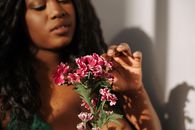 Crop attractive African American female with long wavy hair touching gentle pink pelargonium flower in bedroom
