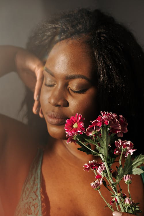 Free Beautiful sensitive African American female touching face with hand and fragrant pink geranium flowers while standing with eyes closed in bedroom Stock Photo
