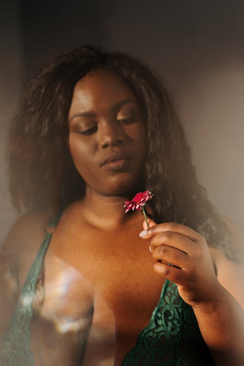 Sensitive plus sized African American female wearing lace lingerie touching and looking at fragrant pink flower in bedroom