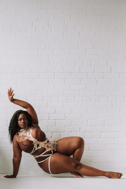 Full body gorgeous plump African American female wearing creative sexy underwear sitting on floor with arm raised graciously and looking at camera