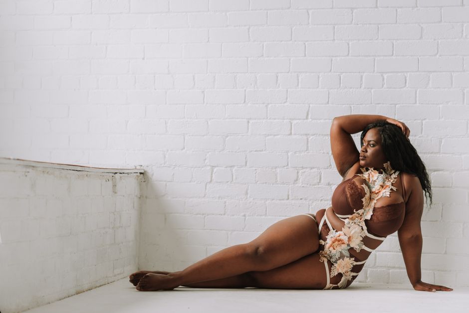 Full length seductive plus sized African American female in underwear touching hair and looking away while lying on floor graciously in light brick studio