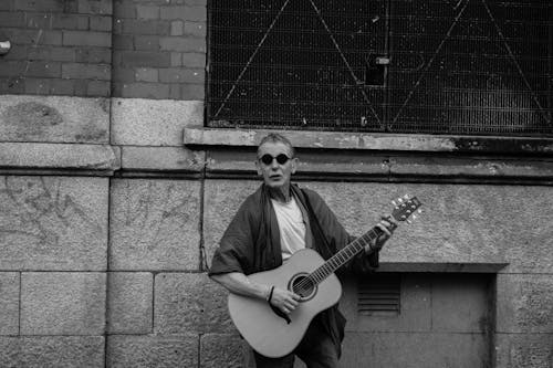 Grayscale Photo of a Man Playing Acoustic Guitar on the Street