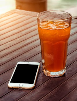 Glass of iced beverage with a smartphone on a wooden outdoor table in bright sunlight.