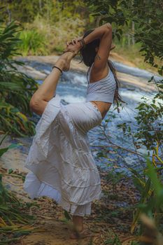 Full body side view of unrecognizable barefoot female standing on one leg while practicing yoga in woodland near flowing stream