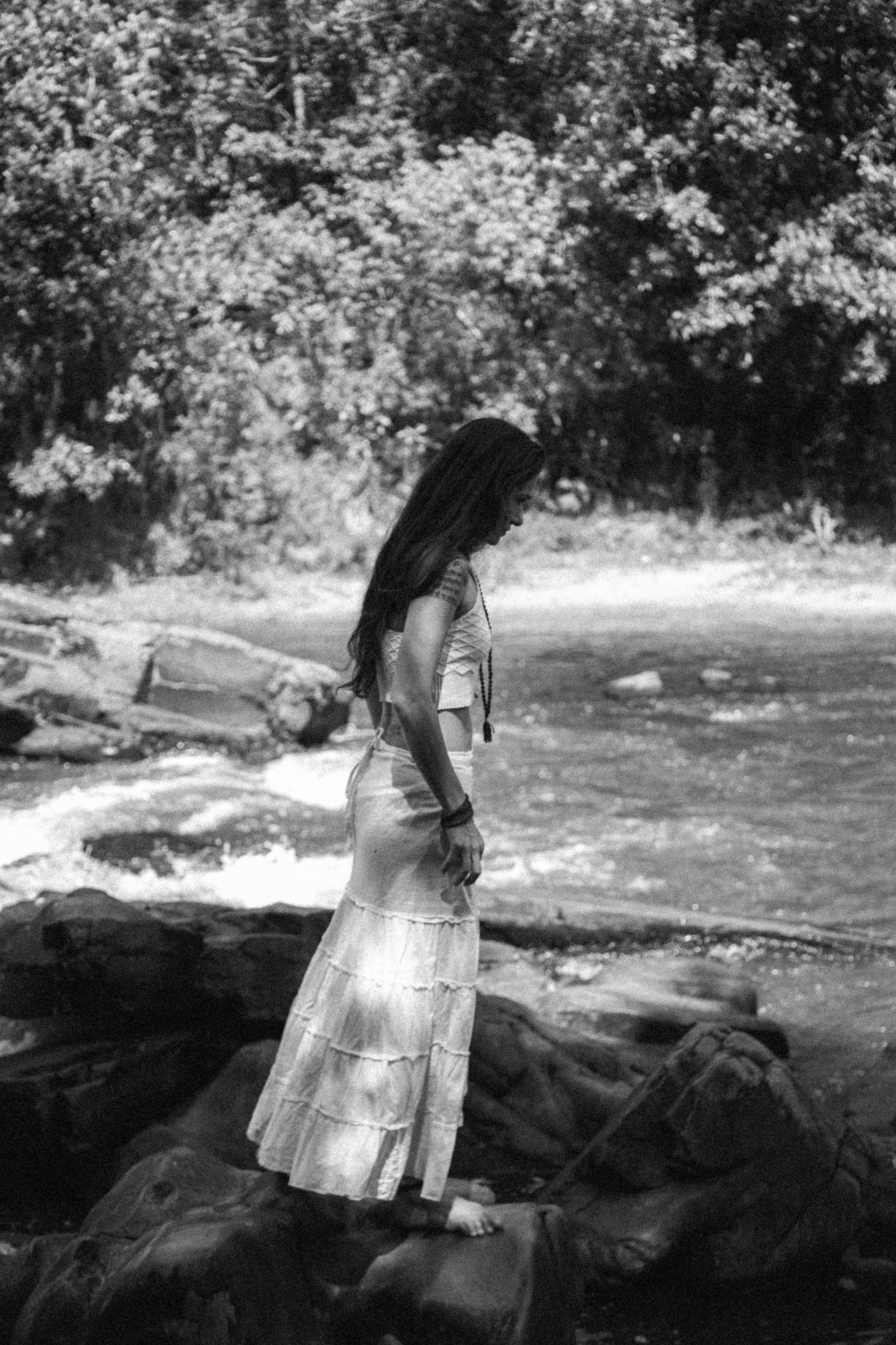 Black and white full body side view of calm female in elegant wear standing on rocky shore near water against forest with trees