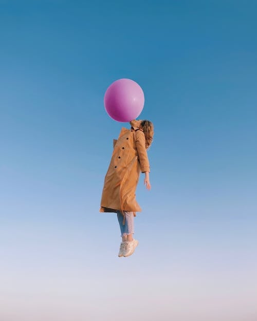 Side view of woman wearing trendy clothes with trench coat flying in sky and blowing enormous balloon