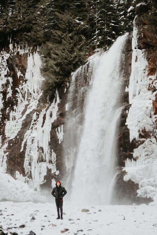 穿著黑色外套和黑色褲子的人站在瀑布附近的積雪覆蓋的地面上