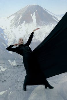 Full body of young enchantress with long white hair in black Gothic dress standing on snowy mountainous terrain with raised arm