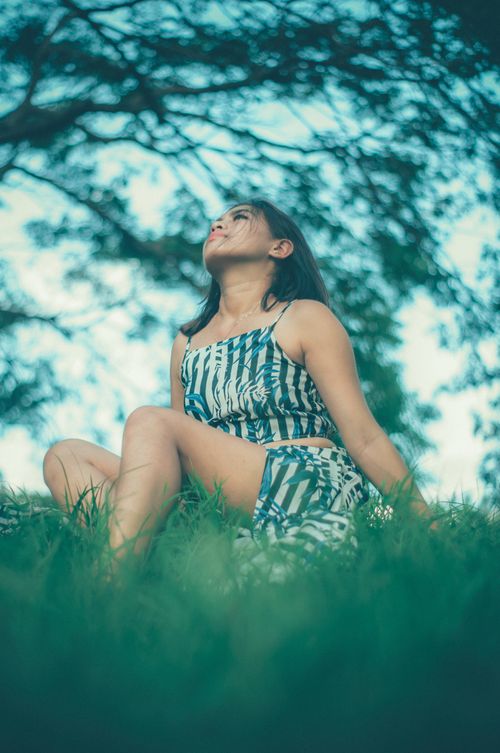 Free Ground level of young female in top and skirt looking up while sitting on green grassy lawn in nature with trees on summer day Stock Photo