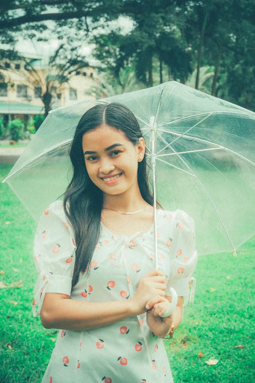 Positive ethnic female with long hair wearing dress standing under umbrella on green lawn and looking at camera