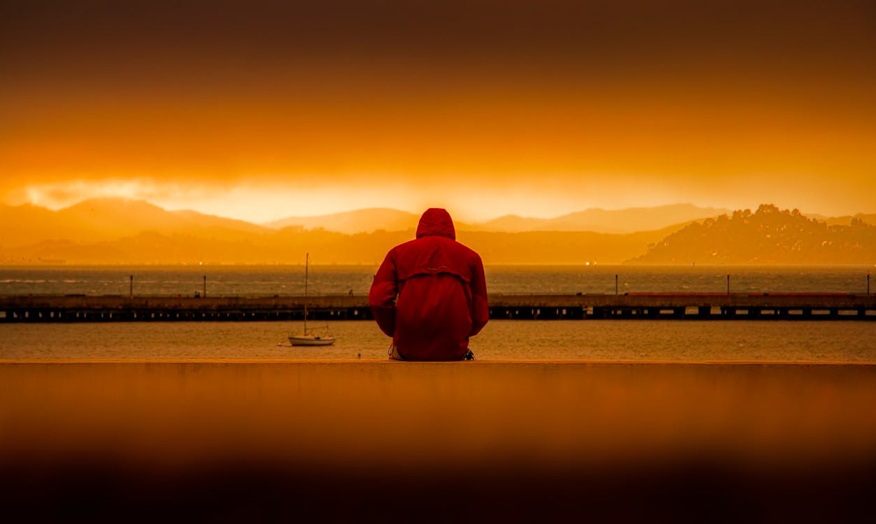 Person Wearing Red Hoodie Sitting In Front of Body of Water