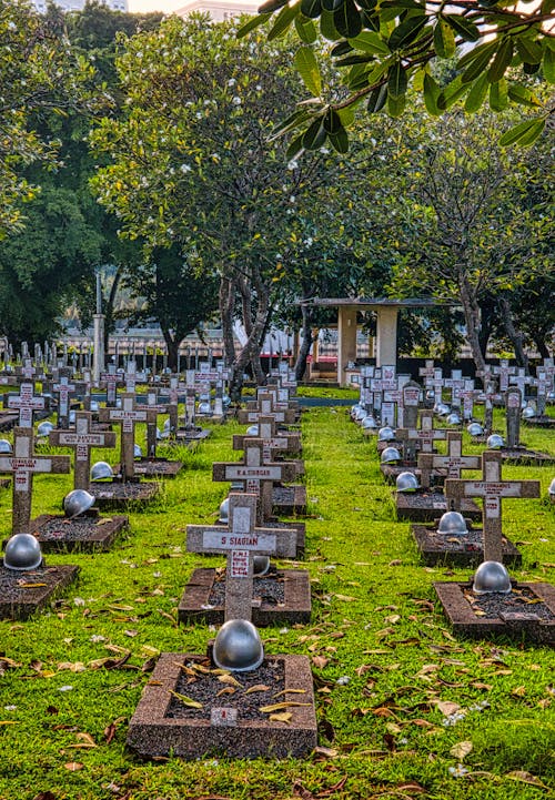 Gravestones with military helmets located on grassy ground in national cemetery with lush tall plants and structure in Kalibata in Indonesia