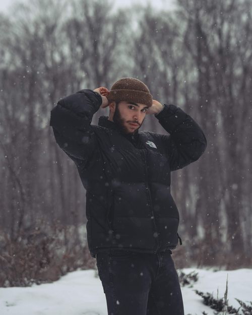 Free A man wearing a jacket and knit cap adjusts his beanie in a snowy forest setting. Stock Photo