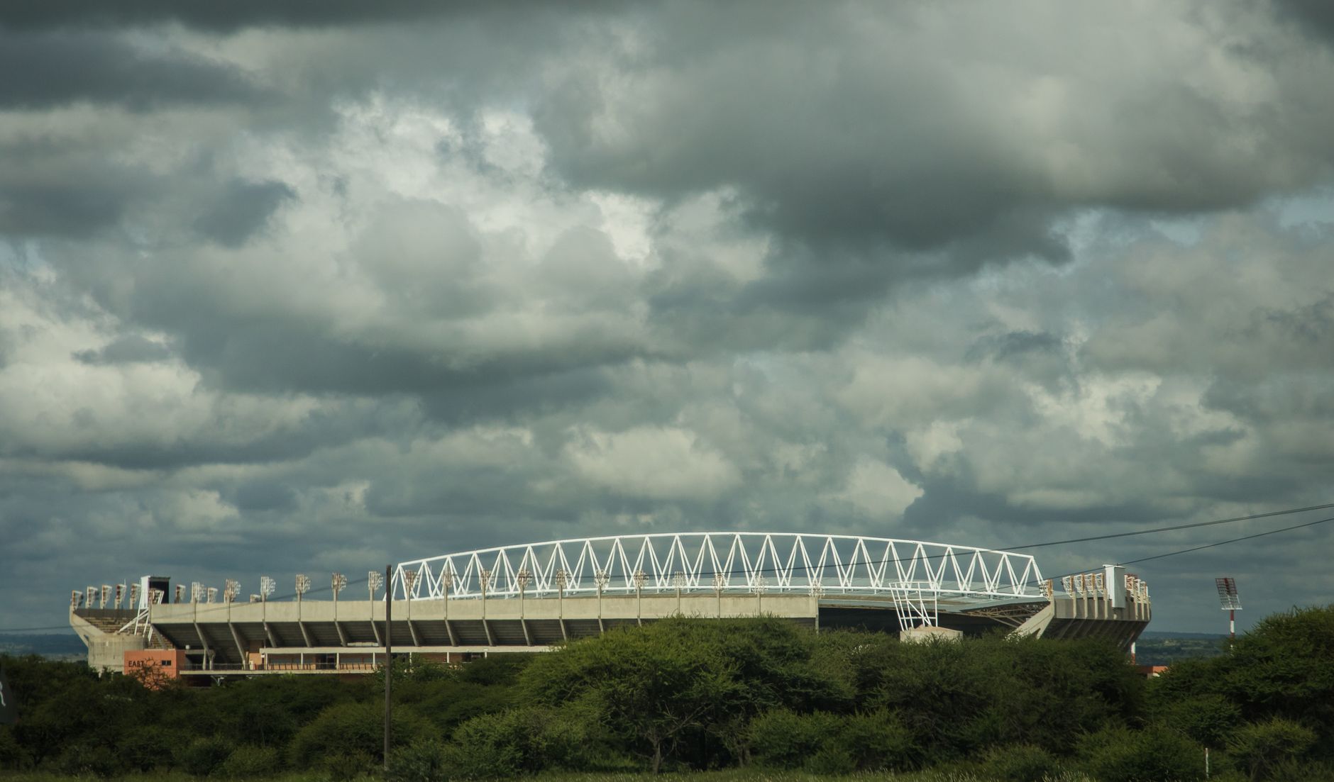 A large modern stadium under a moody cloudy sky with greenery in the foreground.