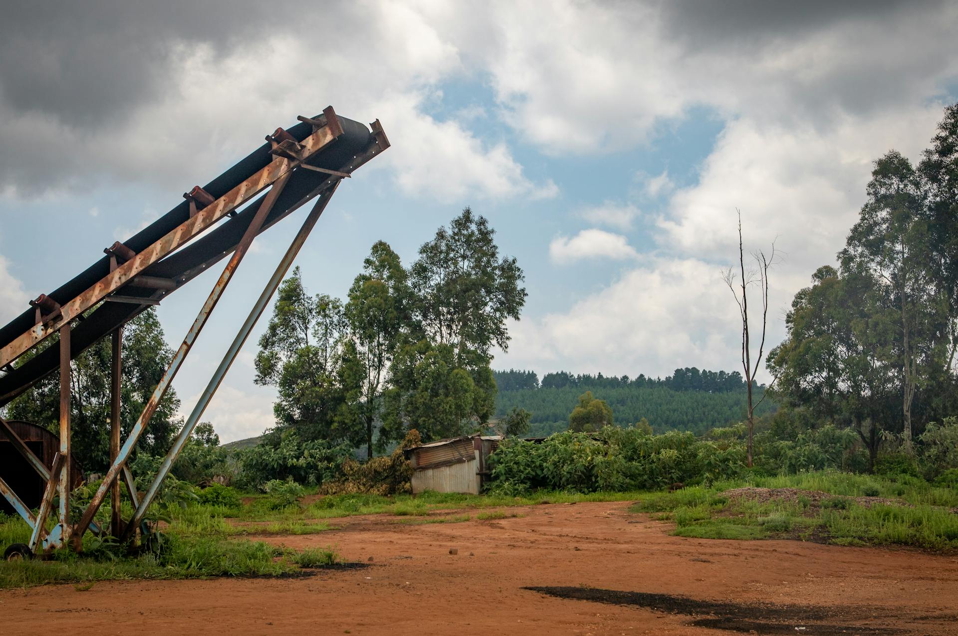 Abandoned conveyor belt amidst rustic rural landscape under cloudy summer sky.