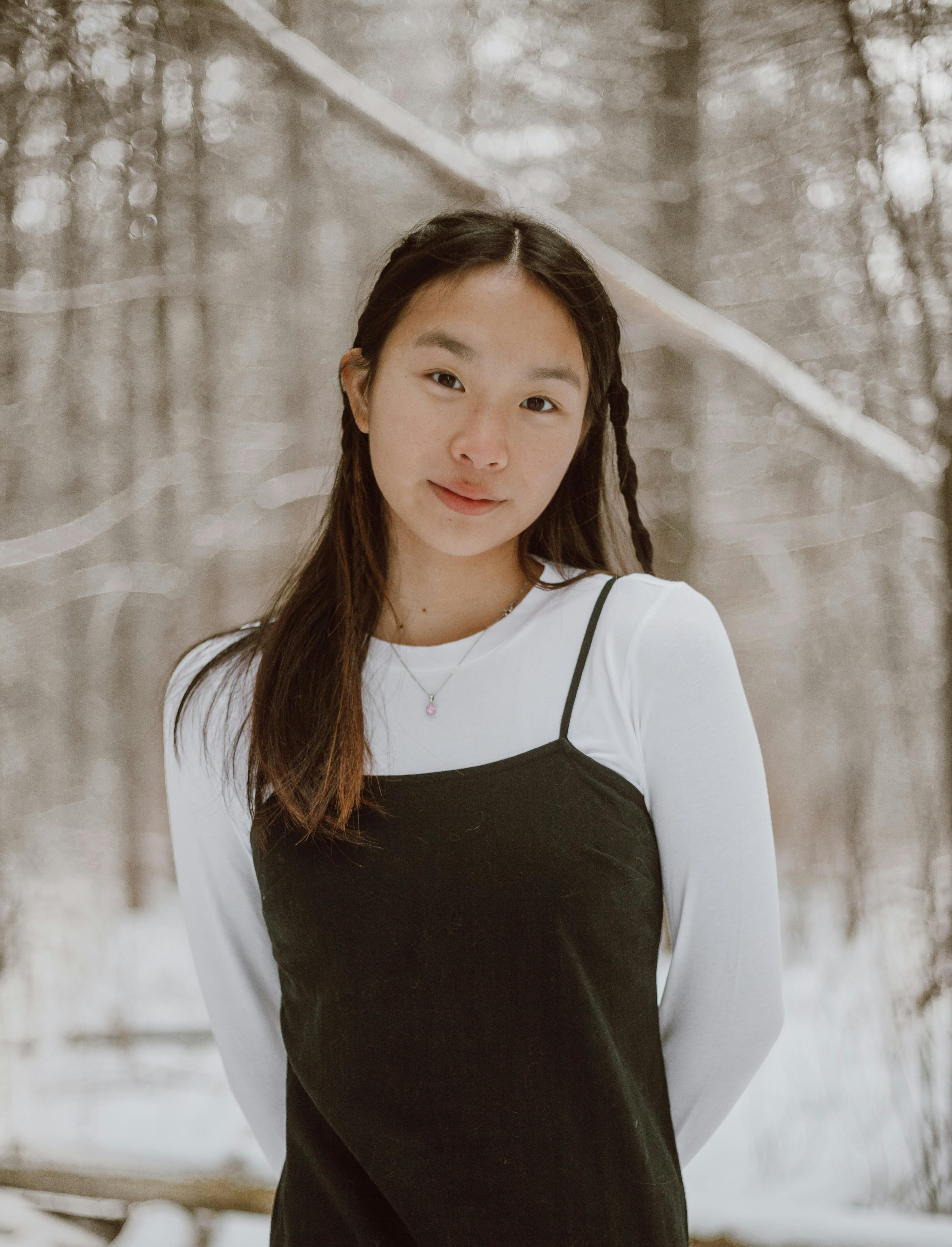 Young gentle ethnic female in casual apparel with hands behind back looking at camera against leafless trees in woods